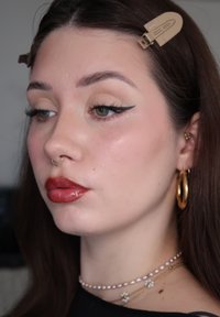 Femme avec un eye-liner ailé, des lèvres rouges brillantes, un anneau de septum en or, des boucles d'oreilles créoles en or, des colliers de perles et de fleurs, et des barrettes beige dans les cheveux.