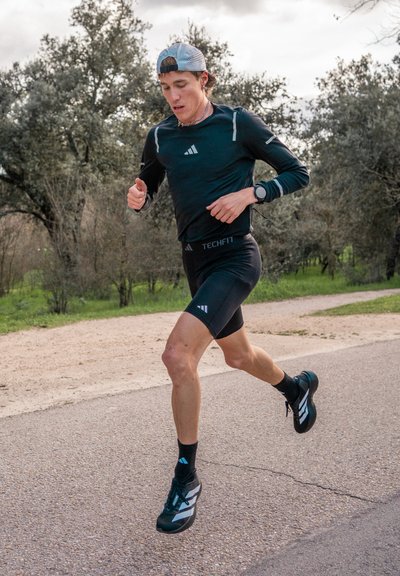 Corredor masculino atlético que lleva una camiseta de manga larga negra, pantalones cortos negros y zapatillas de running en blanco y negro, corriendo por un sendero rodeado de árboles.