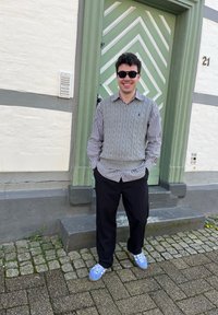 Hallitusest rõngastega vest halli tooniga triibulise särkidega, mustad püksid ja sinised tossud. Seismine rohelise chevron-mustriga ukse ees.