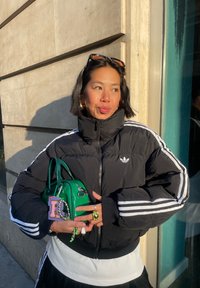 Femme aux cheveux courts et foncés portant une veste Adidas noire et tenant un sac à main vert avec un porte-clés décoratif, debout près d'un mur en pierre sous la lumière du soleil.