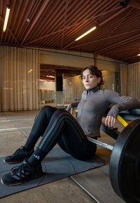 Fitted grey zip-up athletic top and black leggings with vertical stripes; black sports shoes; barbell in gym setting; textured flooring.