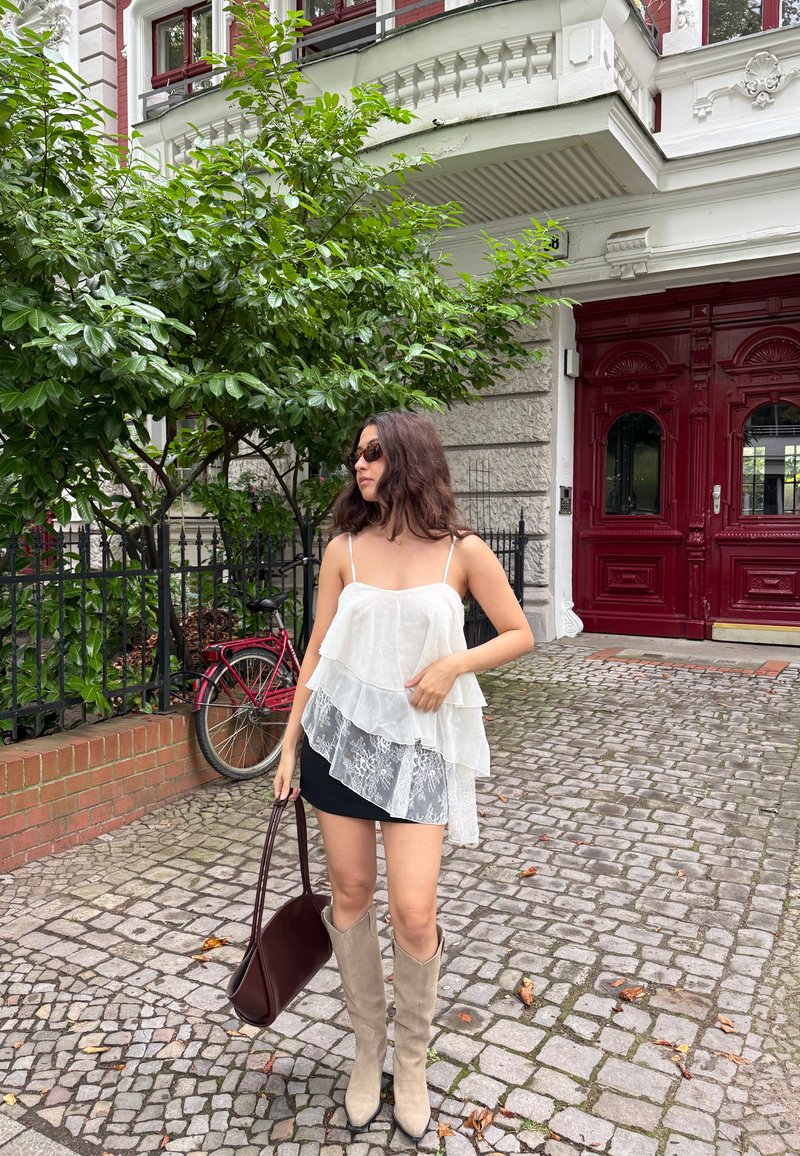 Femme portant des lunettes de soleil, un haut blanc à couches, une jupe noire et des bottes beiges, tenant un sac à main foncé, debout sur un trottoir pavé près d'une porte rouge et d'un arbre vert.