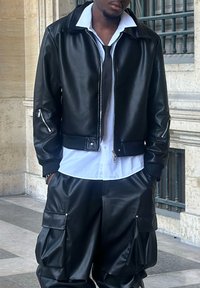 Black leather bomber jacket with a collar and zippered accents, worn over a white shirt. Paired with loose-fitting black leather cargo pants.
