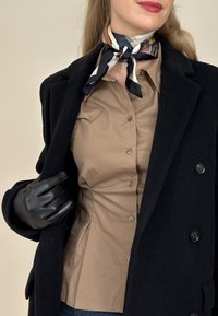 Γυναίκα που φοράει μαύρο παλτό, μαύρο δερμάτινο γάντι, μπεζ πουκάμισο με κουμπιά και μεταξωτό φουλάρι με σχέδιο δεμένο γύρω από το λαιμό.