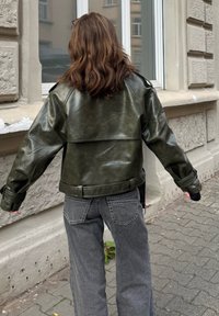 Kožená bunda olivově zelené barvy s krátkým střihem, pásem v pase a širokými rukávy. Kombinována se šedými džíny s širokými nohavicemi. Venkovní prostředí.