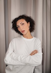 Žena s krátkymi kučeravými vlasmi a kruhovými náušnicami má na sebe bielu košeľu s dlhým rukávom a stojí so založenými rukami pred béžovou záclonou.