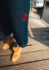 Jeans s kratkim krojem, temno modre barve, z rdečim vezenim logotipom blizu roba, v kombinaciji z rumenimi čevlji.