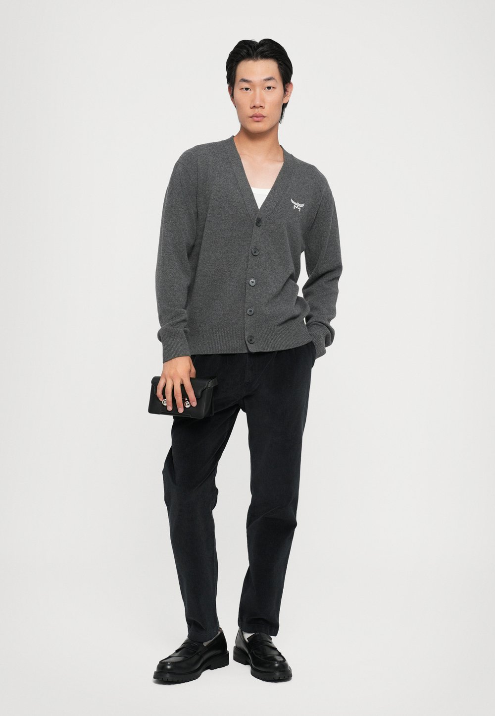 Cardigan gris tricoté avec un col en V et six boutons, associé à un pantalon sombre. Le modèle tient une pochette noire. Design simple et épuré.