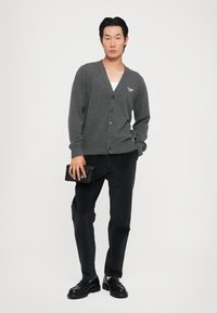Cardigan gris tricoté avec un col en V et six boutons, associé à un pantalon sombre. Le modèle tient une pochette noire. Design simple et épuré.