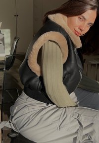 Schwarze Lederweste mit beigem Kunstfellkragen; gerippter olivgrüner Ärmel blitzt hervor; Model sitzt vor einem hellen Hintergrund.