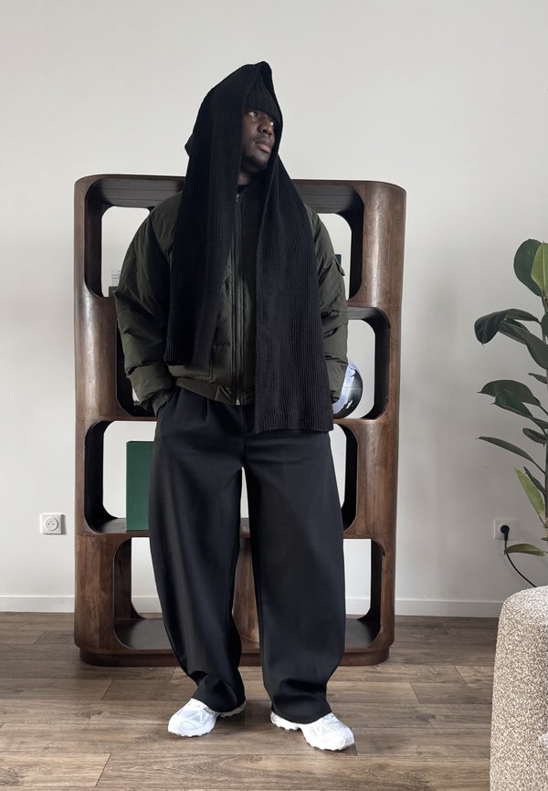 Mann trägt übergroße schwarze Hose, grüne Bomberjacke, weiße Turnschuhe und einen langen schwarzen Schal, der über seinen Kopf drapiert ist, in einem modernen Raum.