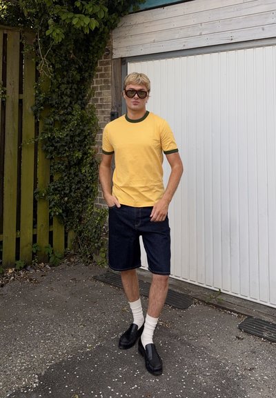 Joven con camiseta amarilla, pantalones cortos oscuros, calcetines blancos, zapatos negros y gafas de sol, de pie al aire libre junto a una puerta de garaje blanca y una valla de madera.