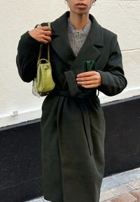 Dark green, long wool coat with a tie belt, wide lapels, and a textured grey sweater underneath. Holding a small green bag and a phone.