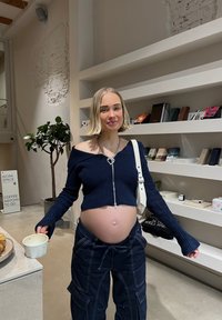 Marineblauwe geribbelde gebreide cropped trui met een rits, in een ontspannen pasvorm, gecombineerd met donkere cargobroeken. Houdt een witte mok vast, staat in een minimalistische ruimte.