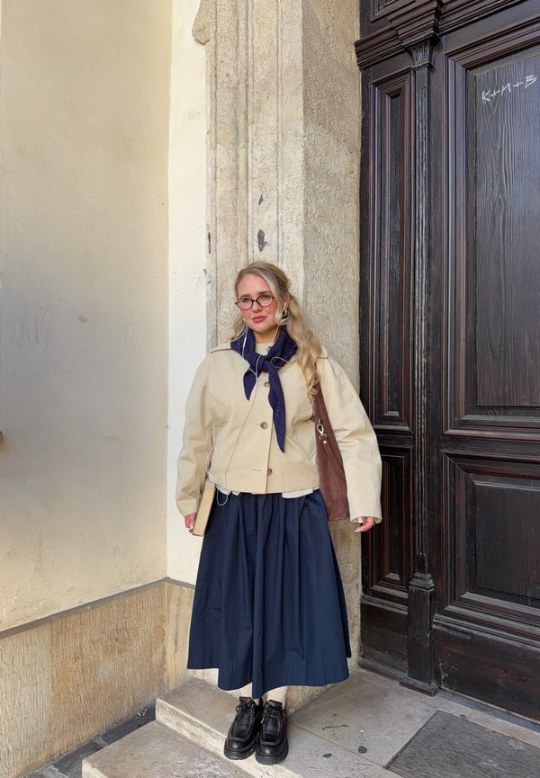 Femme avec lunettes et longs cheveux blonds, debout contre un mur de pierre près d'une grande porte en bois sombre, portant une veste beige et une jupe marine.