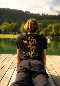 Čierne tričko s farebným kvetinovým dizajnom a textom na chrbte. Nositeľ sedí na drevenej platforme pri vodnej ploche.