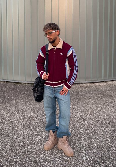 Suéter de punto granate con rayas beige y azul, jeans azul claro y botas gruesas beige. Bolsa negra colgada del hombro, gafas en la cara.