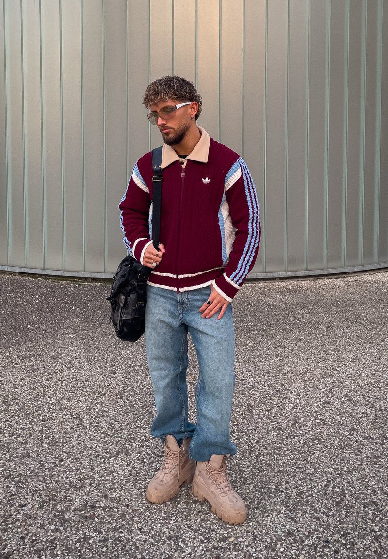 Maglione lavorato a maglia color amaranto con strisce beige e blu, jeans azzurri chiari e stivali beige robusti. Borsa nera a tracolla, occhiali sul viso.