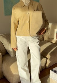 Casaco bege de camurça com colarinho e fecho de botão, combinado com calças largas brancas; segurando uma mala bege combinando.