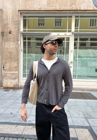 Cardigan grigio con bottoni, t-shirt bianca, pantaloni neri e borsa a tracolla beige; indossando un berretto mimetico e occhiali da sole, in piedi all'aperto.