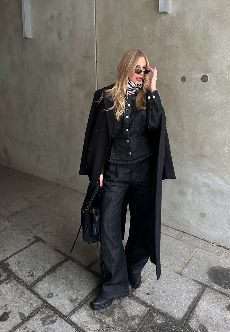 Femme ajustant ses lunettes de soleil, portant un manteau noir sur un costume sombre et une écharpe imprimée zèbre, debout sur un trottoir carrelé contre un mur gris.