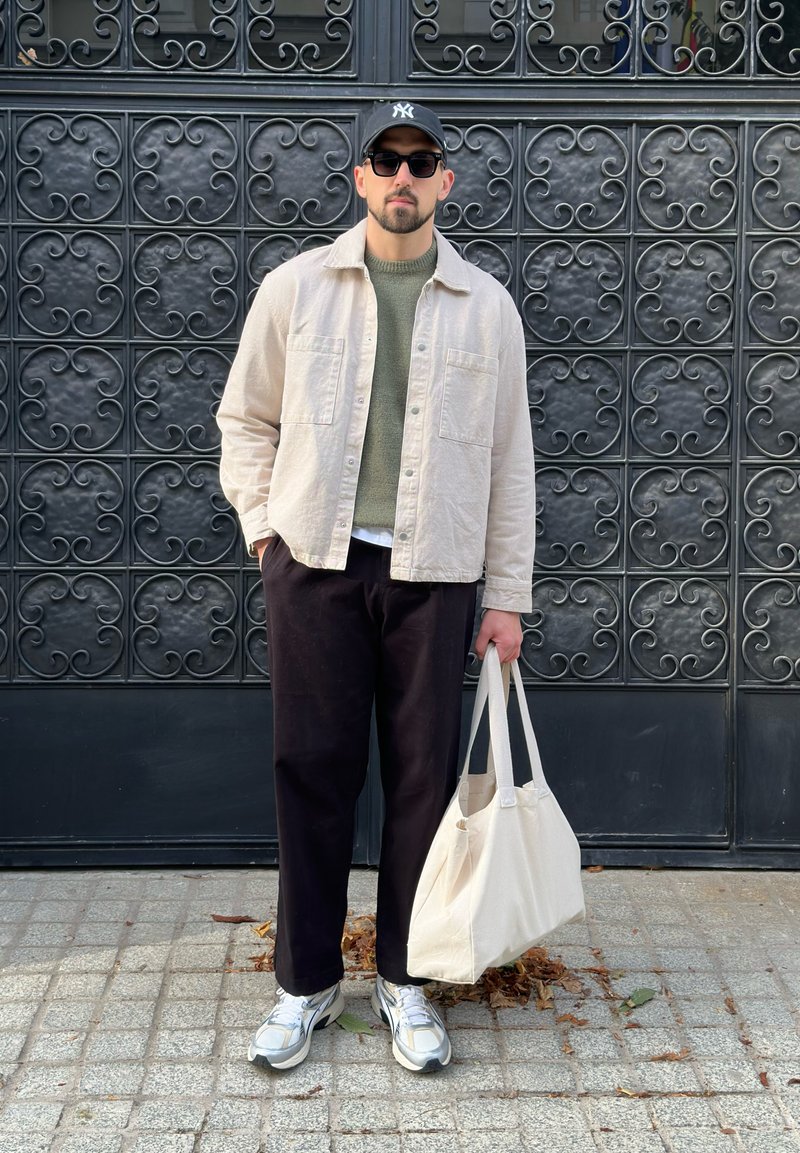Uomo che indossa un berretto nero, occhiali da sole, giacca leggera, maglione verde, pantaloni neri, sneakers bianche, mentre tiene una grande borsa bianca vicino a un cancello metallico decorato.