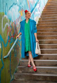 Μια γυναίκα με μπλε παλτό πάνω από ένα πράσινο τρυπητό φόρεμα στέκεται σε σκυρόδεμα σκαλοπάτια, φορώντας κόκκινα παπούτσια και κρατώντας μια λευκή τσάντα.