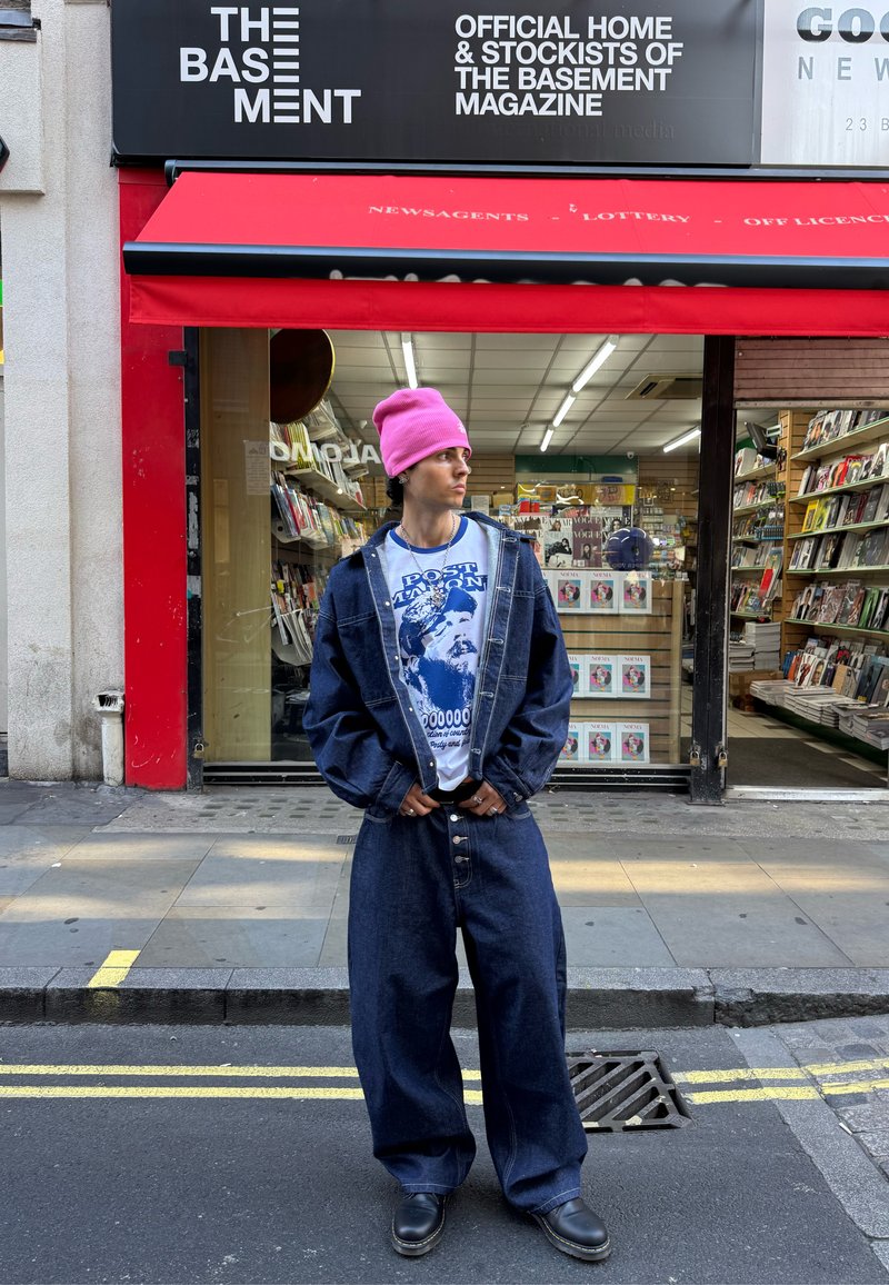 Jeune personne portant un bonnet rose, une veste en jean surdimensionnée et un jean, debout dans la rue devant un magasin de magazines avec un auvent rouge.