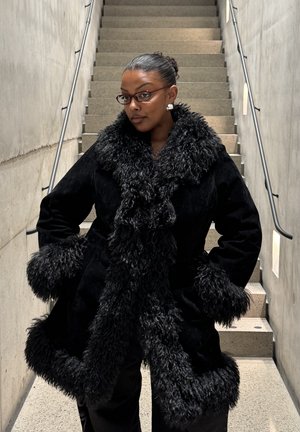 Persona con gafas posa con confianza en unas escaleras interiores, vistiendo un abrigo negro con un gran cuello y puños de pelo esponjoso, en un entorno minimalista de concreto.