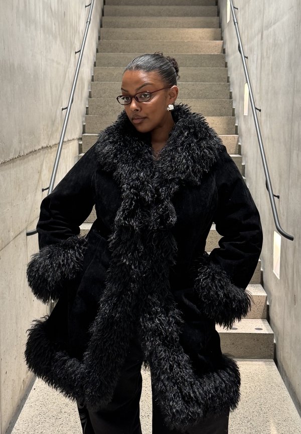 Person med briller står selvsikkert på indendørs trapper og bærer sort frakke med stor, fluffy krave og manchetter i en minimalistisk betonindretning.