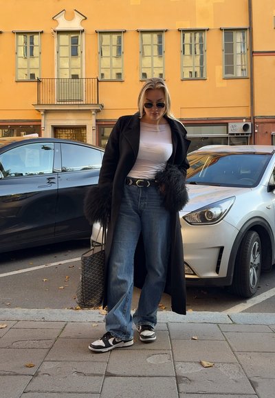 Abrigo largo negro con puños de piel, camisa blanca ceñida, jeans de tiro alto azules y zapatillas en blanco y negro. Bolsa de tejido al lado.