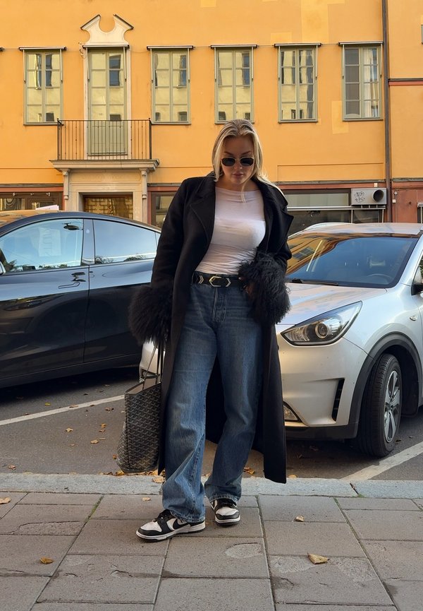 Zwarte lange jas met bontmanchetten, witte getailleerde blouse, hooggerande blauwe jeans en zwart-witte sneakers. Geweven tas aan de zijkant.