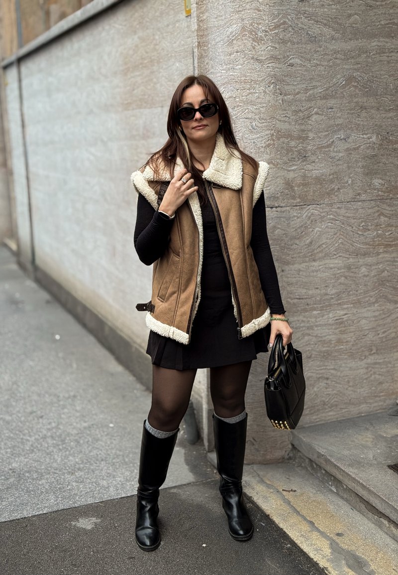 Femme portant des lunettes de soleil, un gilet en peau retournée marron, une robe noire, des collants noirs et des bottes noires montant jusqu'aux genoux, tenant un sac à main noir sur un trottoir urbain.