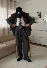 Costume gris avec une chemise blanche et une cravate rayée, châle marron drapé sur les épaules, casquette noire et chaussures noires. Pièce lumineuse avec mobilier beige.