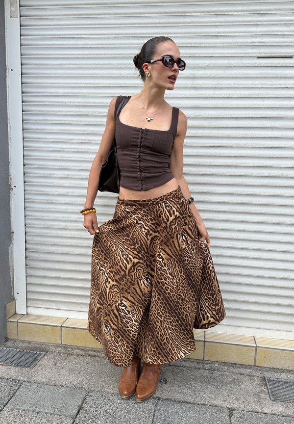 Femme portant des lunettes de soleil, un haut marron sans manches, une jupe large à imprimé léopard et des bottes marron, debout devant une porte à volet blanche.