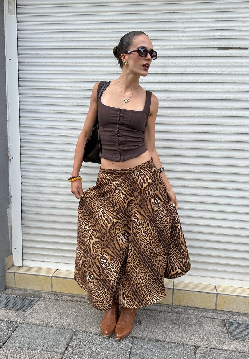 Femme portant des lunettes de soleil, un haut marron sans manches, une jupe large à imprimé léopard et des bottes marron, debout devant une porte à volet blanche.
