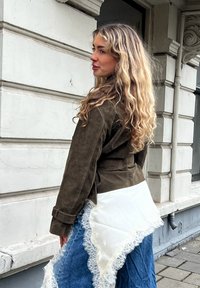 Femme aux longs cheveux blonds ondulés portant une veste marron, un jean bleu et une écharpe blanche bordée de dentelle attachée autour de la taille, debout près d'un bâtiment.