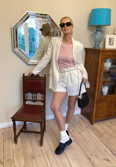 Blazer blanco sobre una blusa rosa, pantalones cortos blancos, zuecos negros y calcetines blancos. Los accesorios incluyen un collar de perlas y un bolso negro.