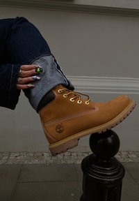 Bottine en cuir marron avec des lacets jaunes, couture blanche et semelle en caoutchouc. L'ourlet du pantalon en denim est retroussé pour révéler la bottine, mettant en valeur les textures superposées.