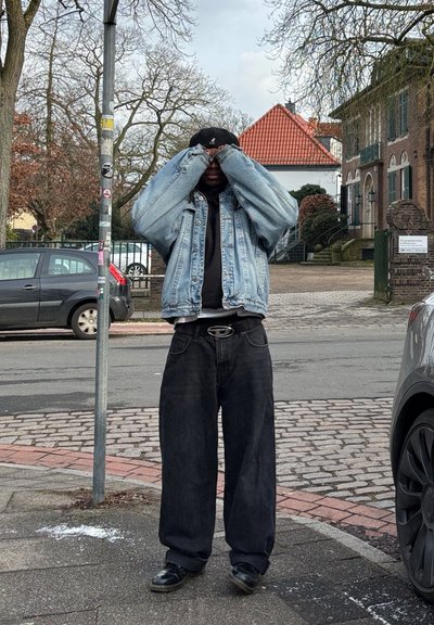 Chaqueta de mezclilla, camisa negra, pantalones negros anchos y zapatos, de pie en una calle con árboles y edificios al fondo.