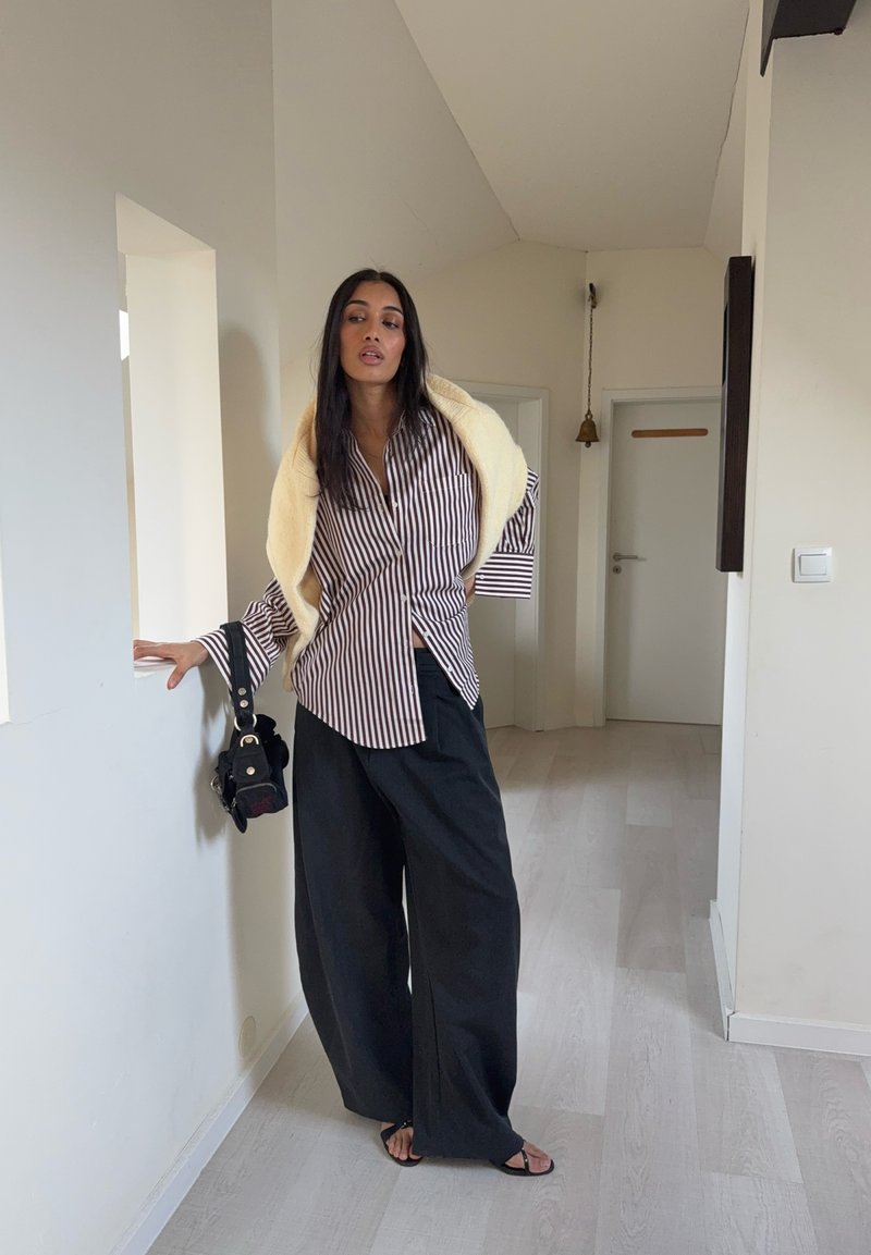 Chemise à rayures boutonnée blanche et bordeaux, pantalon large noir, et châle beige. Sac bandoulière noir avec détails métalliques.