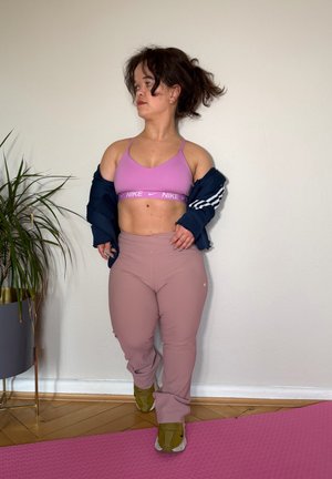 Joven mujer con top deportivo rosa y leggings ajustando una chaqueta azul marino, de pie sobre una esterilla de ejercicio rosa junto a una planta en maceta contra una pared lisa.