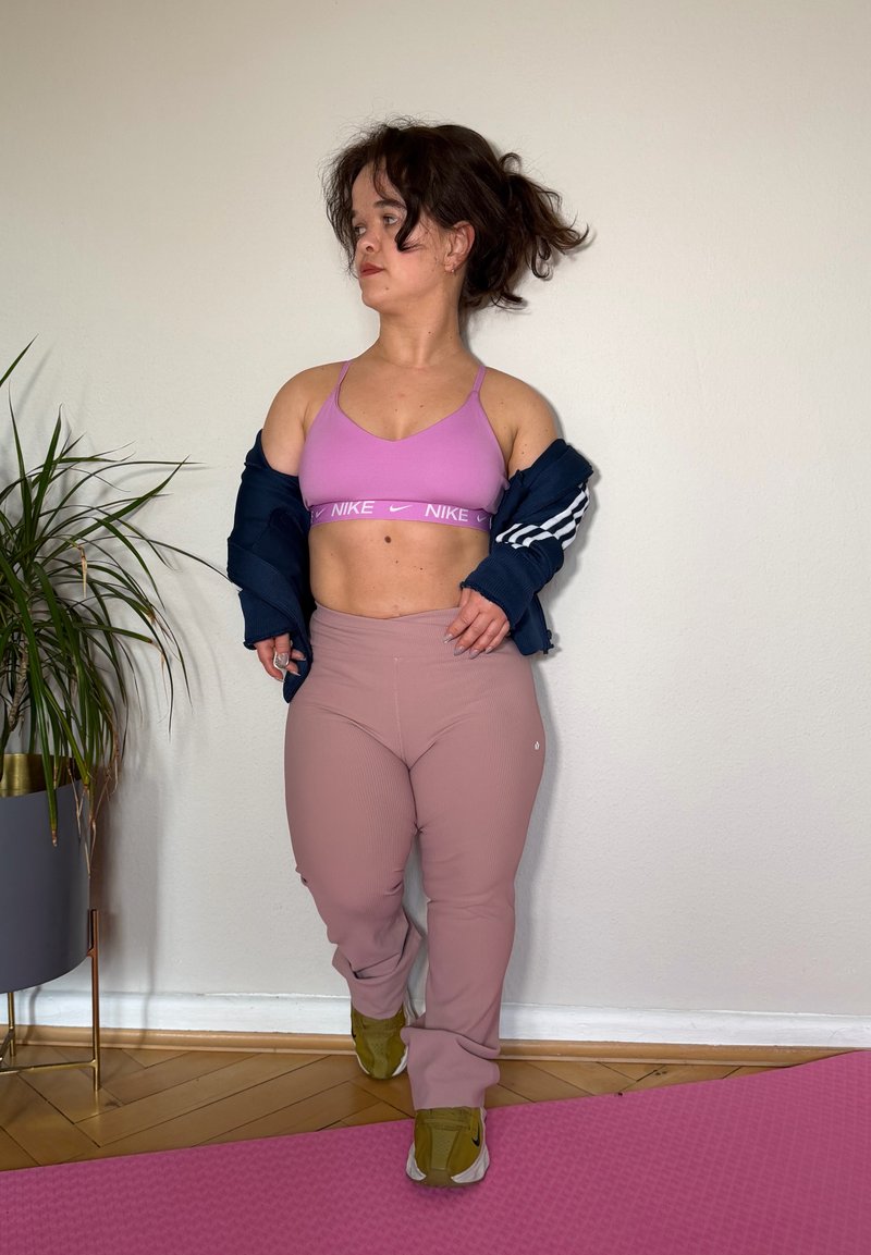 Jeune femme portant un soutien-gorge de sport rose et des leggings, ajustant une veste bleue marine, debout sur un tapis de sport rose à côté d'une plante en pot devant un mur uni.