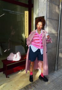 Różowy płaszcz trencz na różowym kardiganie w paski, biała koszula i szorty w prążki; do tego różowe skarpetki i buty na platformie. Trzyma kubek.