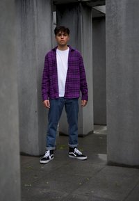 Cămașă flanelată lila cu carouri peste un tricou alb, blugi, și teniși negri cu glezna înaltă, stând între coloane de beton gri.