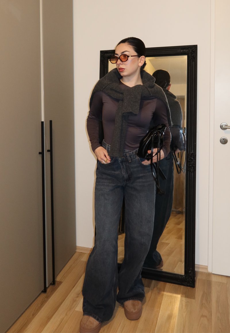 Femme portant des lunettes de soleil foncées, un haut marron à manches longues, un pull gris drapé sur les épaules, un jean à jambes larges, des chaussures beige, tenant un sac noir à l'intérieur.