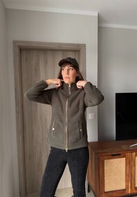 Γυναίκα με πράσινο καπέλο και πράσινο φλις μπουφάν με φερμουάρ, ρυθμίζει το γιακά της, στέκεται μέσα σε ένα δωμάτιο κοντά σε ξύλινη πόρτα και ντουλάπι με τηλεόραση πάνω.