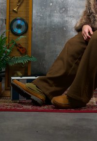 Personne portant un pantalon large marron et des mocassins couleur tan assise sur un tapis à motifs à côté d'une plante et d'une vitrine en bois contenant des instruments de musique.