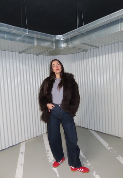 Mujer con abrigo de piel oscuro, camisa gris, pantalones vaqueros anchos oscuros y zapatillas rojas, de pie en un espacio industrial minimalista con paredes onduladas y conductos en el techo.