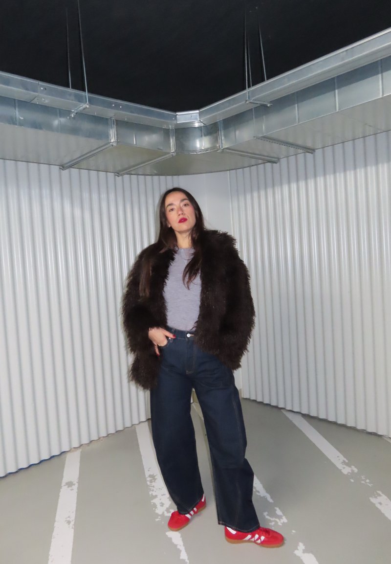 Frau in dunklem Pelzmantel, grauem Hemd, weitem dunklem Jeans und roten Turnschuhen, die in einem minimalistischen Industriegebäude mit Wellblechwänden und Deckenlüftungen steht.